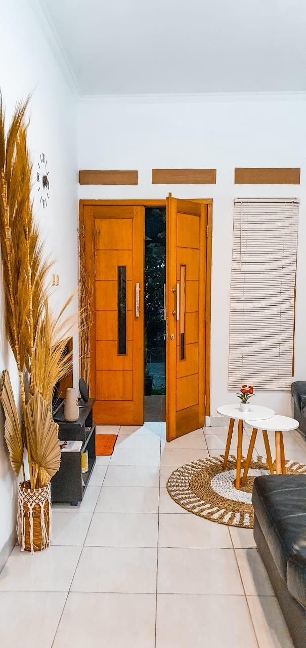 A welcoming entrance features double wooden doors that open to a softly lit living area. A small round coffee table is placed on a circular rug, accompanied by a modern black sofa. A tall decorative plant adds a touch of greenery to the space.