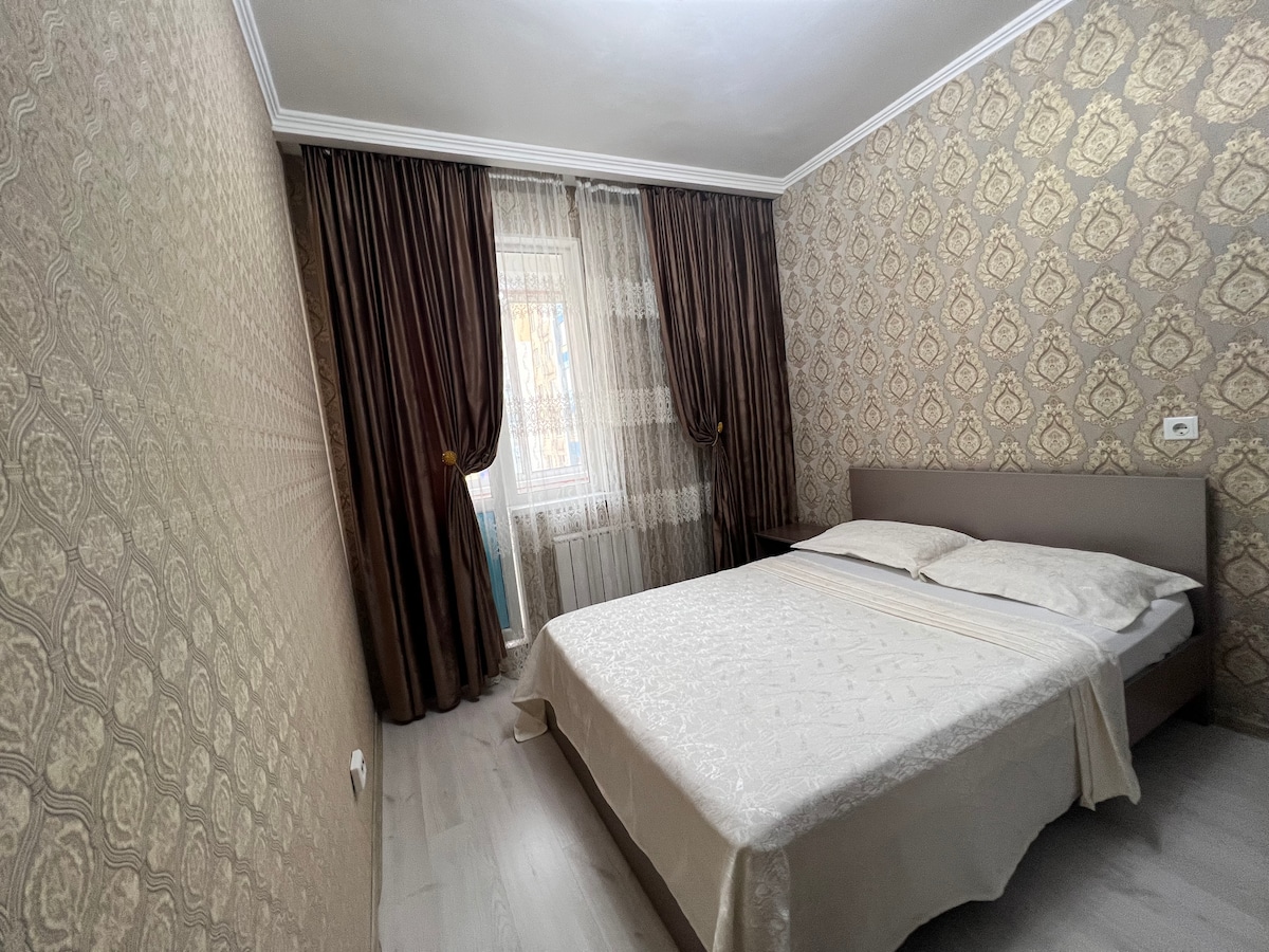 A well-appointed bedroom features a spacious bed dressed in elegant white linens. Soft, neutral-toned wallpaper complements the natural light streaming through the sheer curtains. Richly colored drapes frame the window, adding warmth to the overall space.