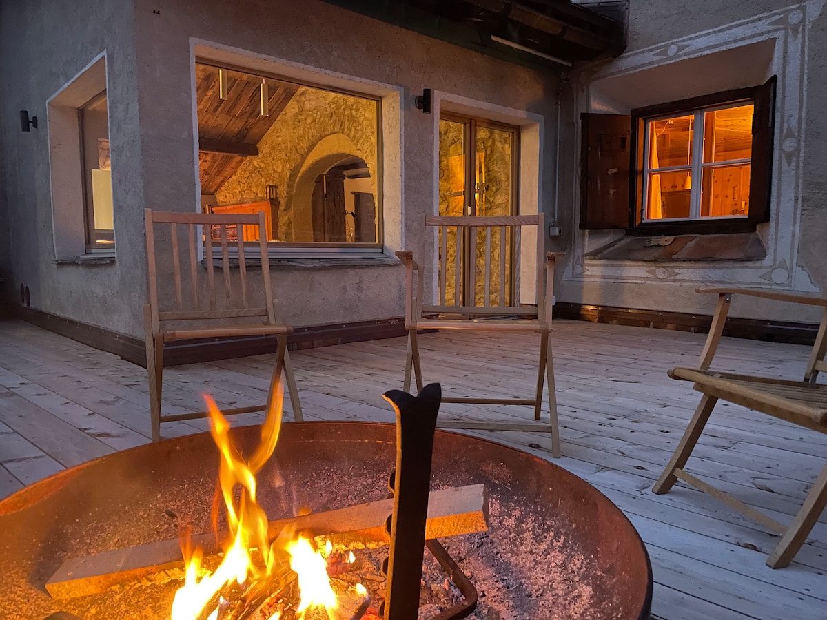 A wooden terrace is featured, with several chairs arranged around a fire pit where flames flicker. The warm glow from windows reflects a welcoming ambiance. The surrounding walls are beautifully crafted stone, evoking a rustic charm.