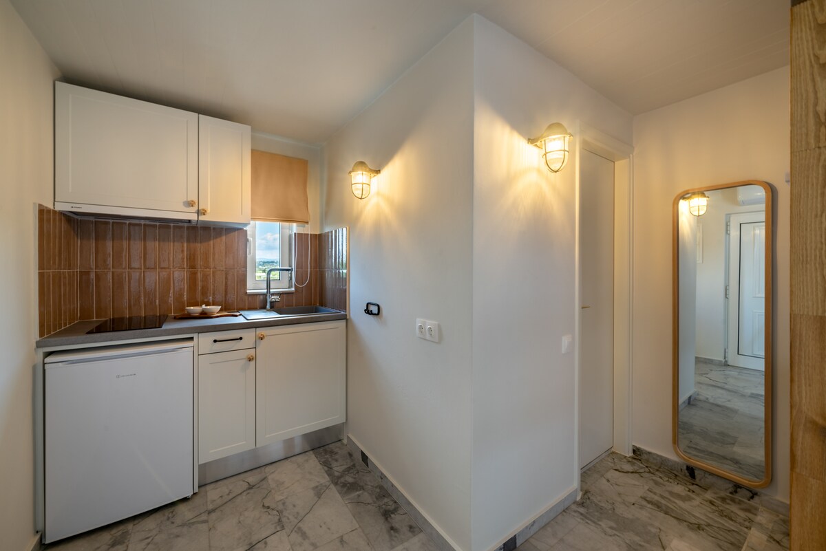 A compact kitchen space features light cabinetry and an integrated fridge. A large mirror reflects the room, while warm lighting is provided by wall-mounted fixtures. The area is complemented by a tiled floor, creating a functional and inviting cooking environment.