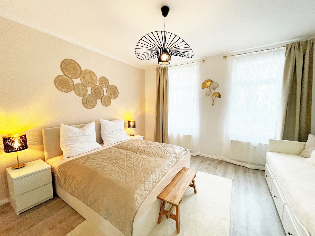 A well-lit bedroom features a spacious bed adorned with neutral bedding. Two bedside lamps provide soft illumination, while decorative wall art made of woven materials adds visual interest. A cozy seating area includes a light-colored sofa, with large windows dressed in simple curtains enhancing the natural light.