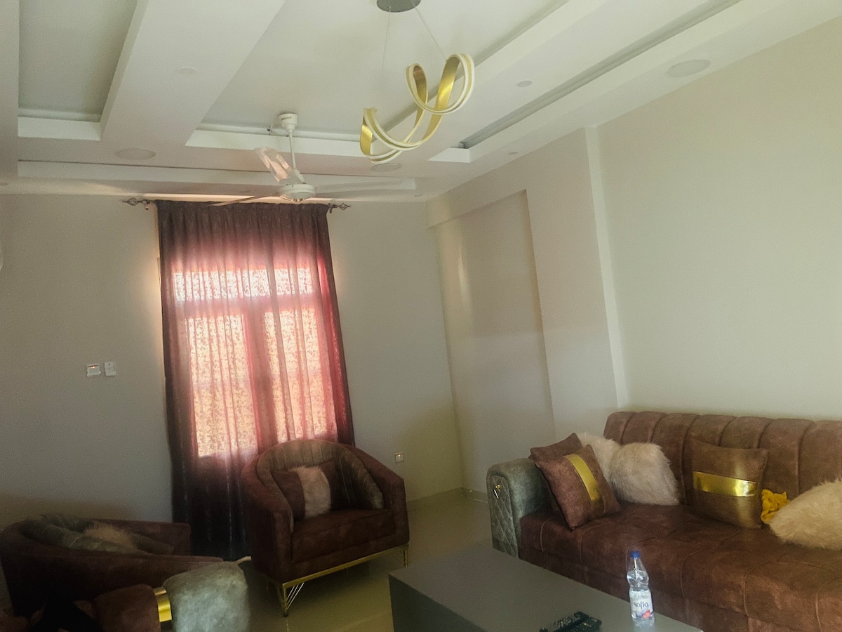 A spacious living area features a combination of plush seating arrangements, including two armchairs and a comfortable sofa. Natural light filters through the large window, which is dressed in rich, dark curtains. A simple ceiling fixture adds modern elegance to the room.