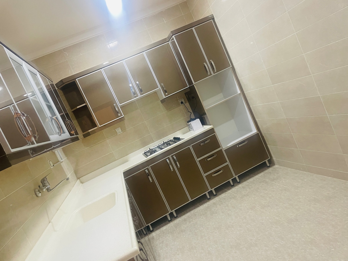 A functional kitchen features modern cabinets with a sleek finish and ample storage. A stove is positioned alongside a countertop, while the sink area is neatly arranged. Neutral-colored tiles create a clean and uncluttered environment, enhancing the kitchen's practicality.