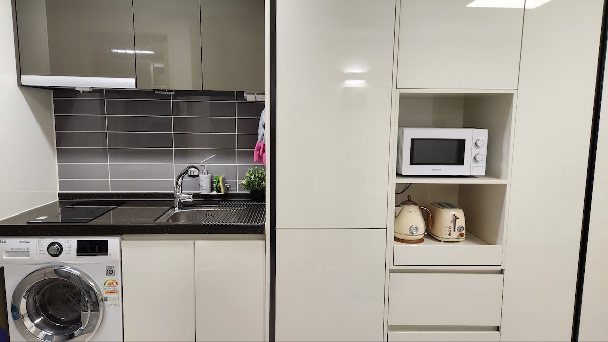 A modern kitchen area features sleek cabinetry and countertops. Essential appliances, including a washing machine, sink, microwave, and kettle, are neatly arranged. Decorative elements, such as a small plant, add a touch of greenery to the contemporary space.