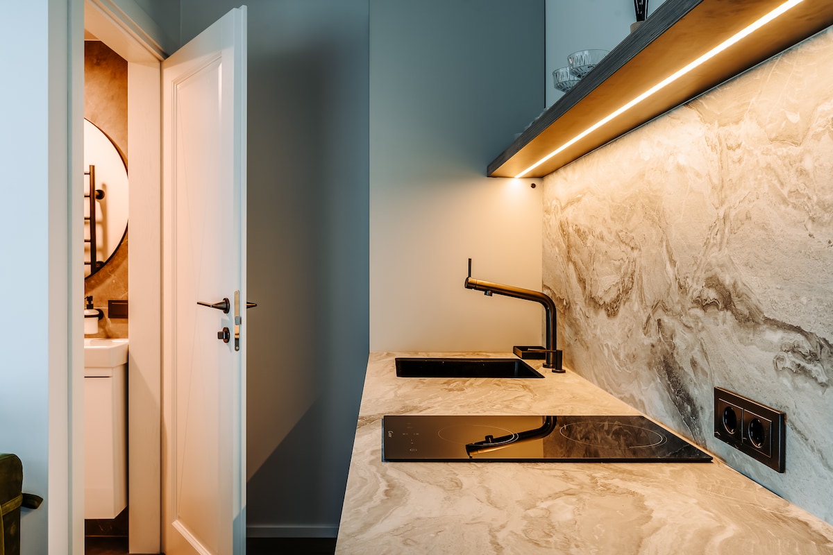 A modern kitchen features a smooth marble countertop with integrated sinks and a sleek cooktop. Under-cabinet lighting highlights the elegant stone surface. A doorway leads to a bathroom area, providing a seamless connection between spaces.