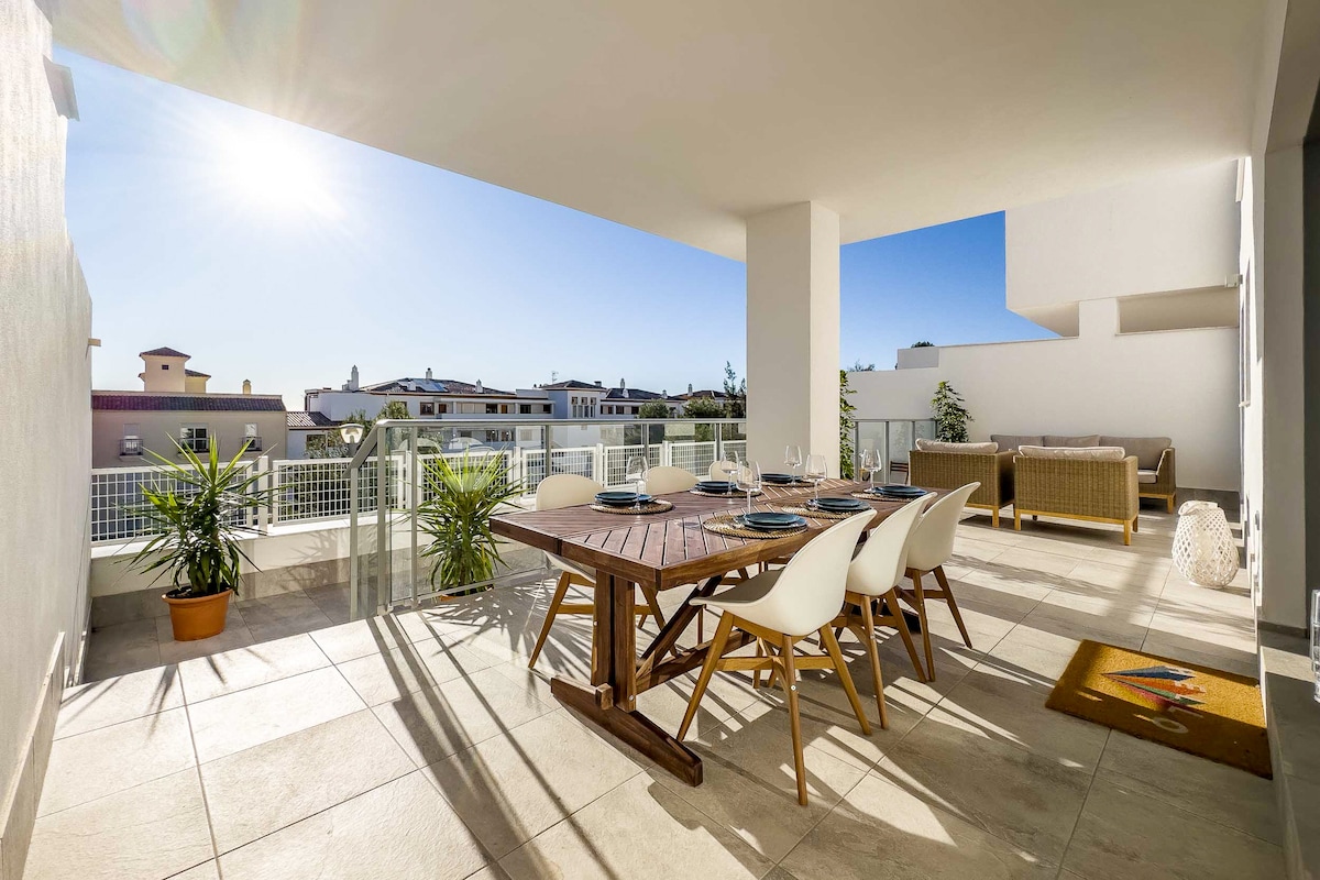 An outdoor terrace features a wooden dining table set for six, complemented by sleek white chairs. Comfortable lounge furniture is arranged near a potted plant, under bright sunlight. The backdrop showcases nearby buildings and a clear blue sky.