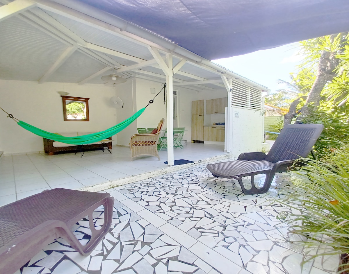 A shaded outdoor patio area features a hammock, two lounge chairs, and a dining table surrounded by tropical plants. The flooring is a distinctive mosaic pattern, and a kitchenette is visible in the background, providing an inviting space for relaxation and meals.