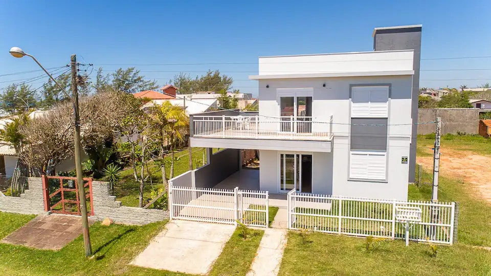 The exterior view of the two-story house features a light-colored facade with multiple windows. A front porch is highlighted, with a railing and outdoor seating available. The surrounding yard is grassy, complemented by landscaping elements and a fenced area, creating a private outdoor space.