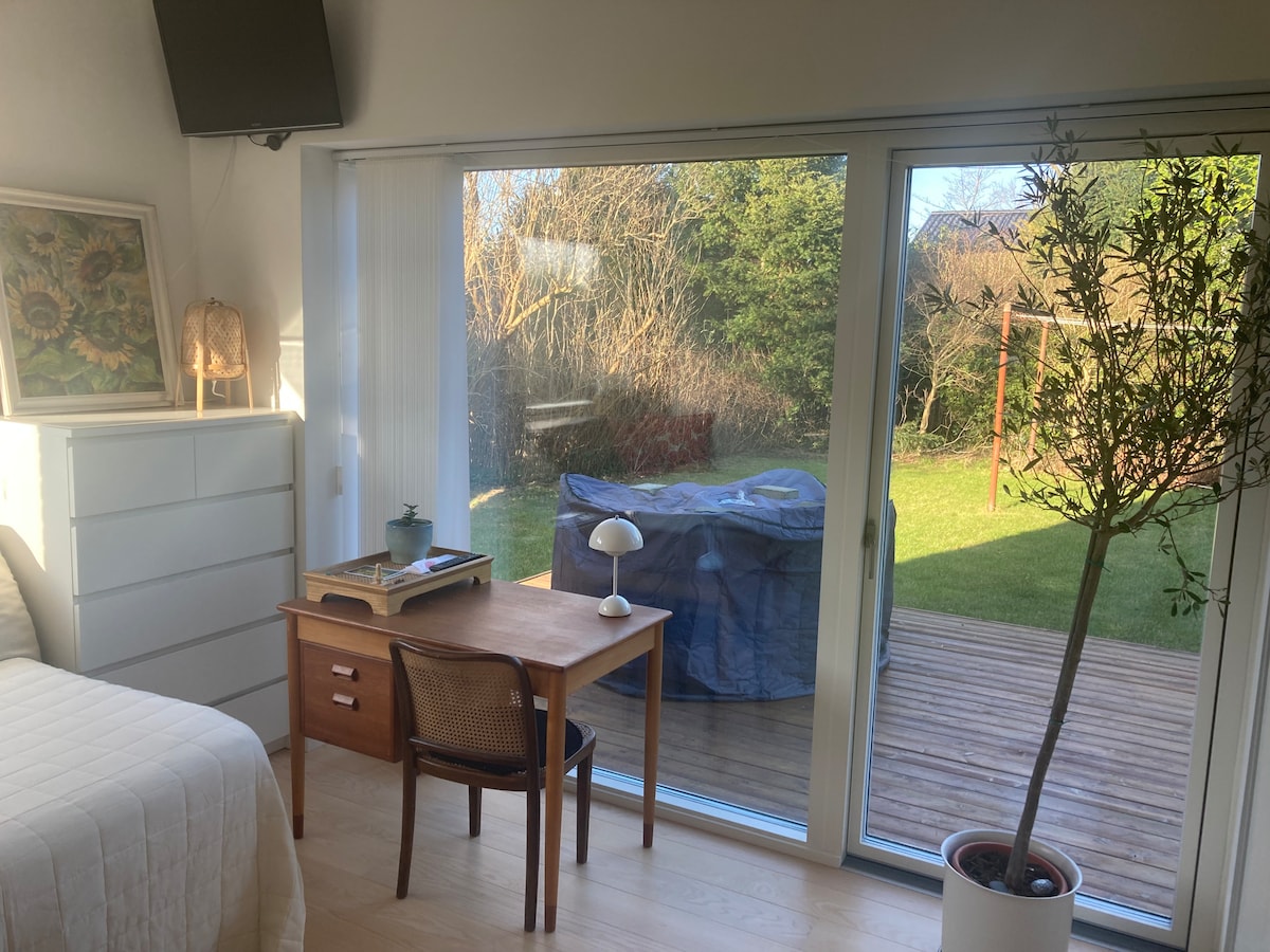 A bright bedroom features large sliding glass doors that showcase a view of a private terrace and garden area. A minimalist wooden desk is positioned beside the window, accompanied by a potted plant. Sunlight softly illuminates the space, creating a serene atmosphere.