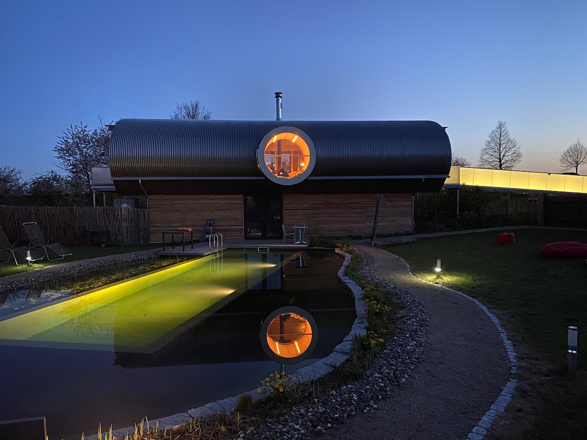 A modern wooden house at twilight showcases a distinctive rounded design with a large circular window. The tranquil swimming pond reflects warm lighting, enhancing the outdoor ambiance. Softly lit pathways and a manicured garden complement the space, inviting a serene evening atmosphere.