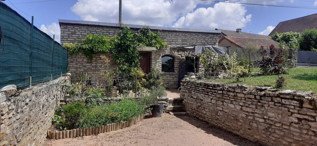 A stone exterior showcases the charming maisonnette surrounded by greenery. A well-tended garden features various plants, while a pathway leads to the entrance. Sunlight illuminates the area, enhancing the tranquil setting of the private property.