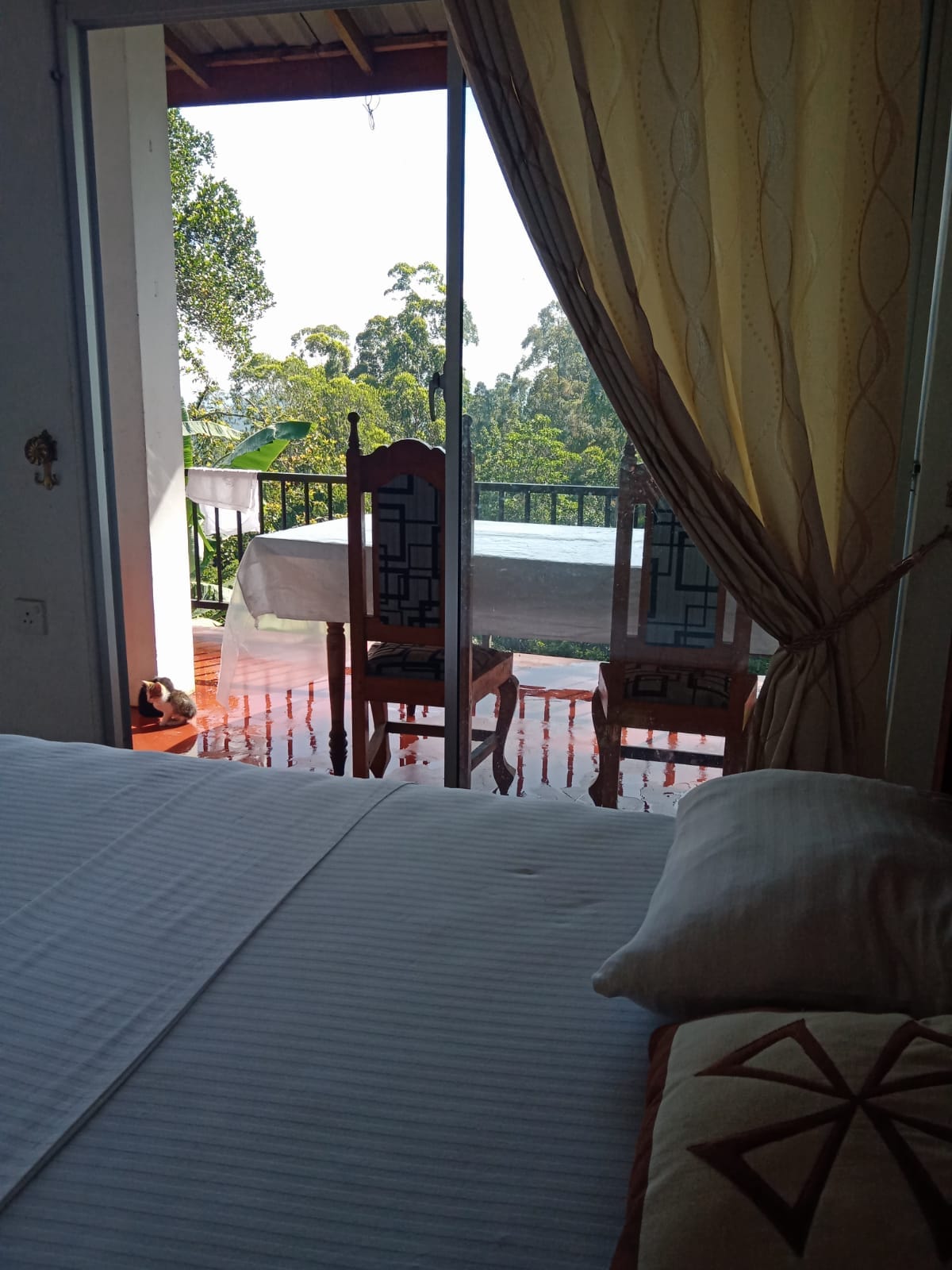 A bedroom features a spacious bed with crisp white linens, complemented by decorative pillows. Large glass doors open to a balcony, where two wooden chairs are positioned. Lush greenery is visible outside, enhancing the connection to nature.