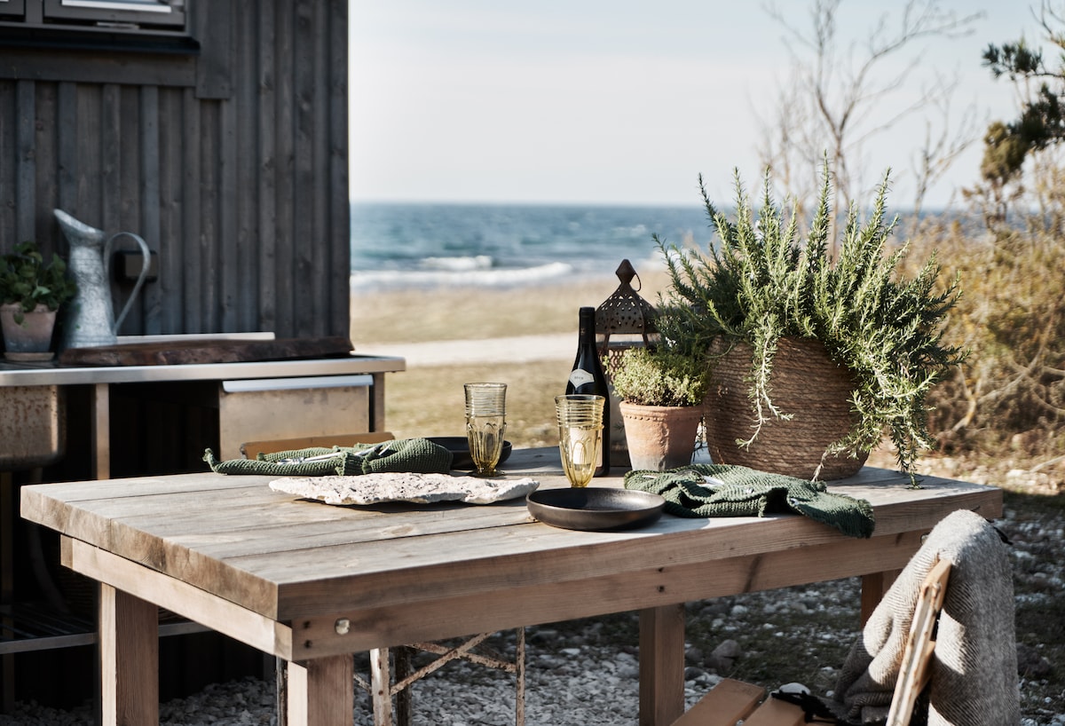 An outdoor dining area features a wooden table set with glasses and a bottle, complemented by a potted plant. The table is positioned with a view of the sea, surrounded by natural landscape.