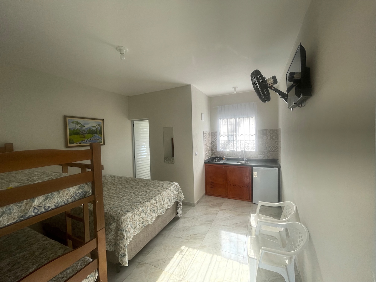 A bright room is furnished with a spacious bed and a bunk bed. A small kitchenette is visible, featuring a refrigerator and cabinets. Two white chairs are positioned near a window that allows natural light to fill the space, highlighting the tiled floor.