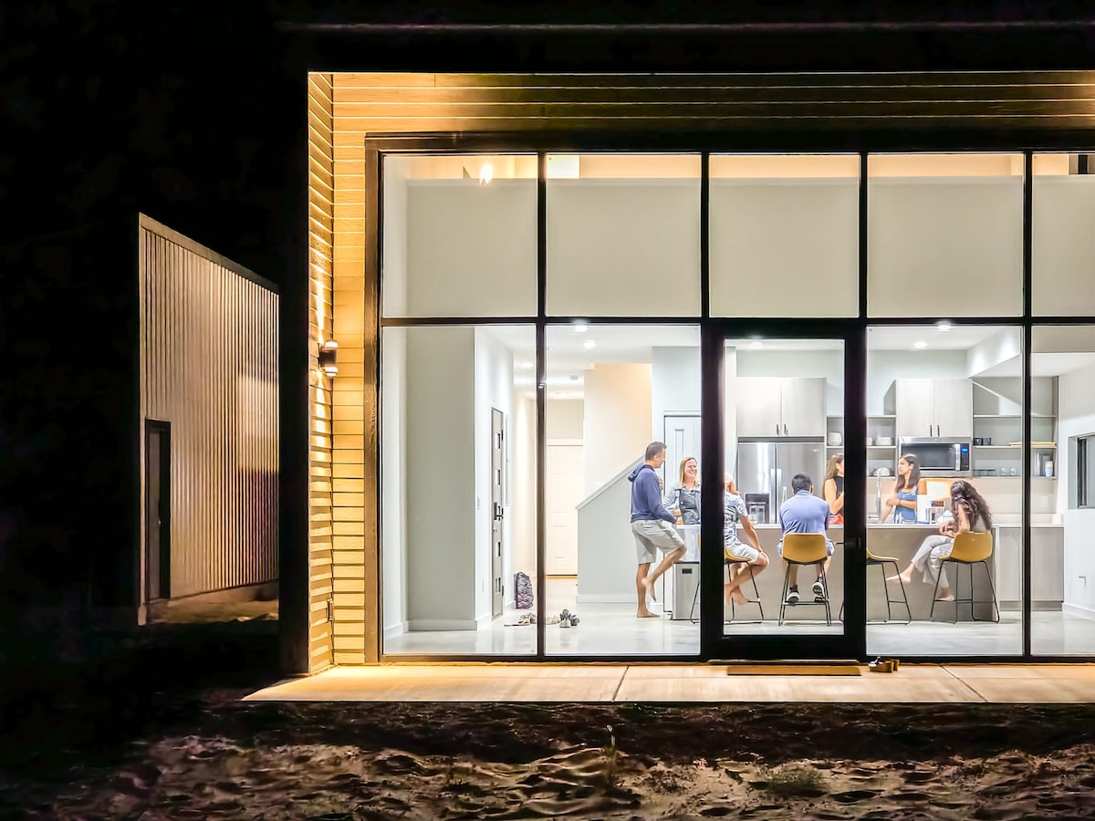 The image shows a modern kitchen and dining area, lit warmly against a dark exterior. Large glass doors provide a view of guests gathered around a table, enjoying a meal together. The kitchen features sleek cabinetry and bright lighting.