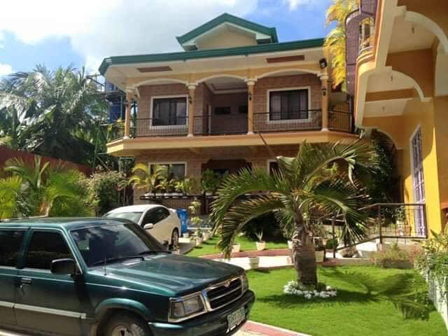 A multi-level building is surrounded by lush greenery, featuring a mix of palm trees and flowering plants. The exterior showcases balconies with black railings, while two parked vehicles are seen in the driveway. The bright sky adds to the overall inviting atmosphere.