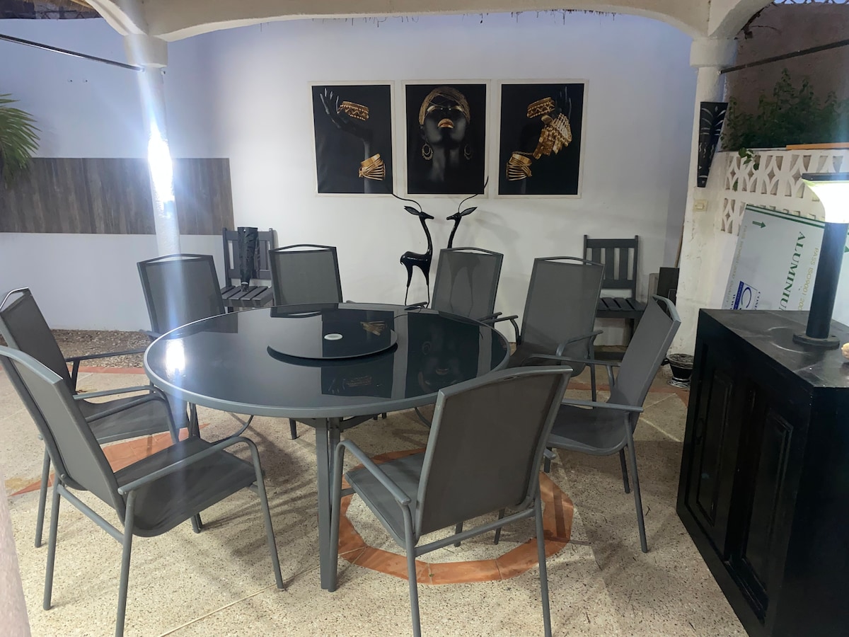 An outdoor dining area features a large circular table surrounded by eight gray chairs. Two artworks are displayed on the wall behind, and a bicycle is positioned nearby. The setting is illuminated by a nearby lamp, enhancing the space's usability at night.