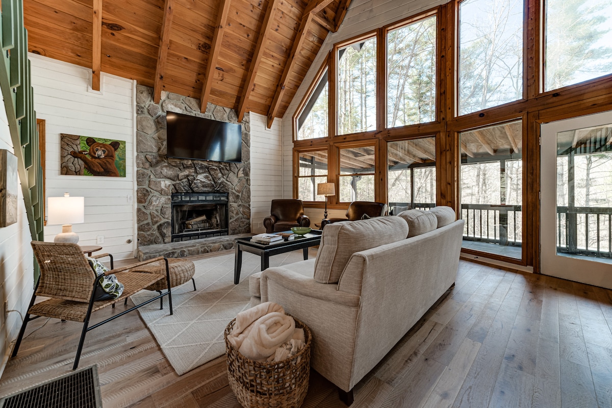 An inviting living area features high ceilings with wooden beams and large windows that allow natural light to fill the space. A stone fireplace is centered against the wall, flanked by comfortable seating. A soft throw is neatly placed on a light-colored sofa.