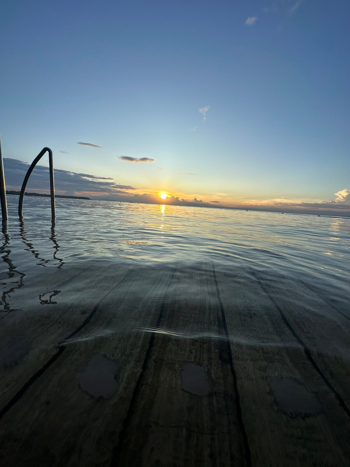 A serene sunset is captured over a calm body of water, reflecting shades of orange and blue. The image includes a pool ladder extending into the water, with wooden decking visible nearby. Soft ripples create a tranquil surface as the day transitions to evening.