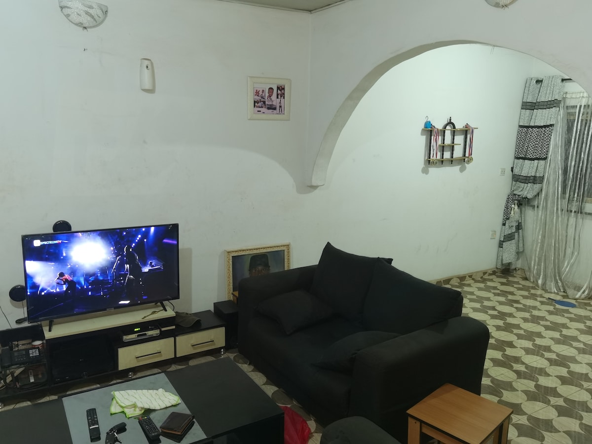 A shared living room is presented, featuring a dark sofa and a low coffee table. A television displays a performance, while patterned flooring covers the space. Light-colored walls and decorative elements, including a shelf and curtains, contribute to the room's functionality.