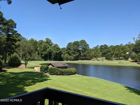A serene view of a lush green lawn extends towards a calm lake. The clear blue sky reflects in the water, while trees line the perimeter, creating a peaceful atmosphere. A small shed is visible near the water's edge, complementing the natural setting.