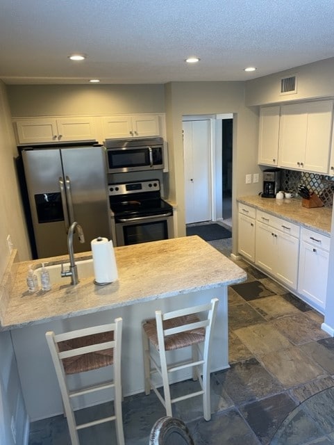 The kitchen features modern stainless steel appliances, including a refrigerator and oven, accompanied by a spacious stone countertop. Light cabinetry complements the tile flooring, and a breakfast bar with two stools provides a casual dining option. The area is well-lit with overhead lighting.