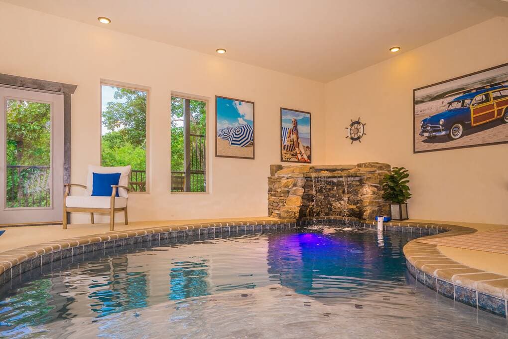 A spacious indoor pool area features a stone waterfall, complemented by decorative wall art. Natural light filters through large windows, illuminating the tranquil water. A comfortable chair is placed nearby, providing a relaxing space to enjoy the serene ambiance.