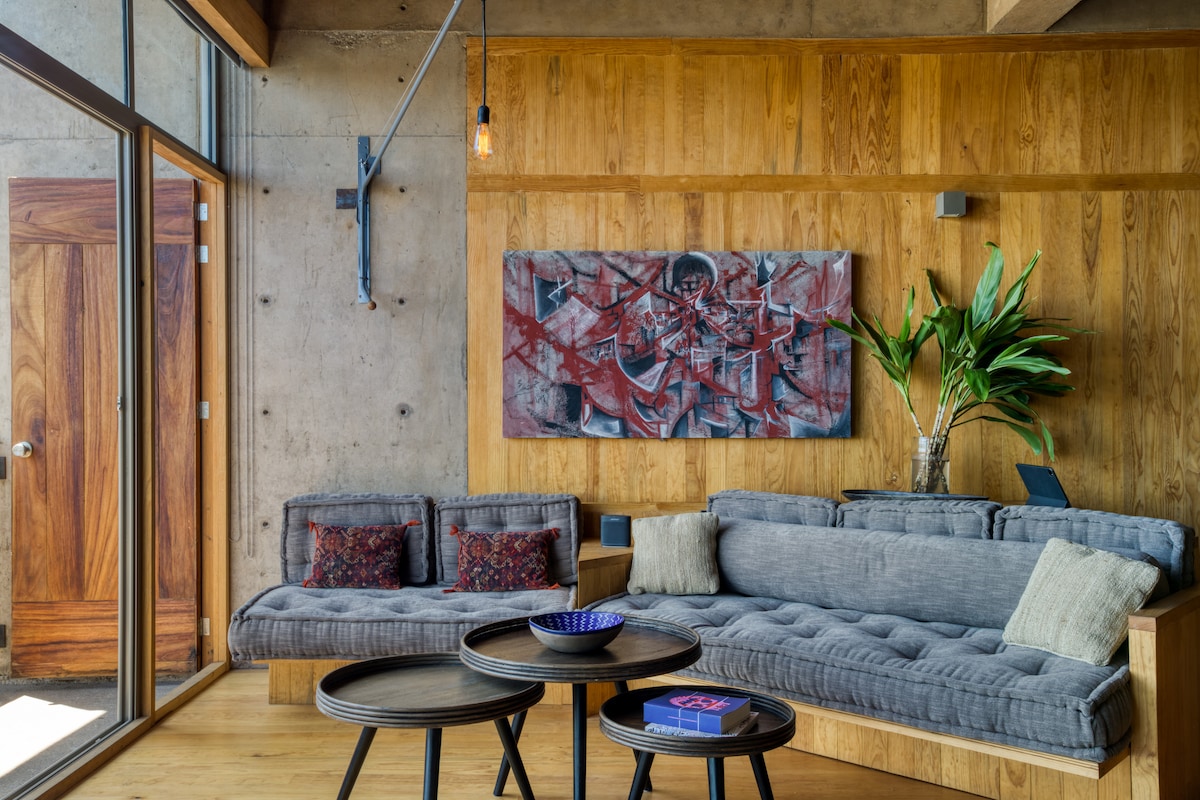 A modern living area features a comfortable grey sectional sofa with accent pillows, positioned next to large glass doors that allow natural light. A piece of abstract art hangs on the wall above, and a plant adds a touch of greenery. Two round tables are placed in front.