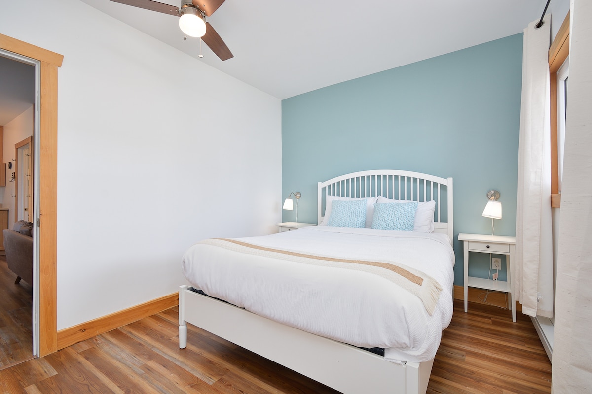A spacious bedroom features a soft bed against a light blue accent wall. Two bedside lamps provide illumination beside the bed. Natural light enters through a nearby window, accompanied by light-colored curtains. A door to another room is visible in the background.