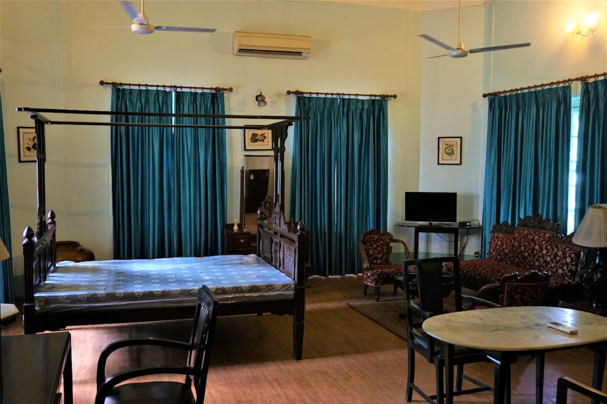 An airy studio apartment is highlighted by a spacious four-poster bed and large windows draped with teal curtains. Antique furniture includes a cozy sitting area with patterned upholstery and a round marble dining table. A modern desk with a TV stands in one corner.