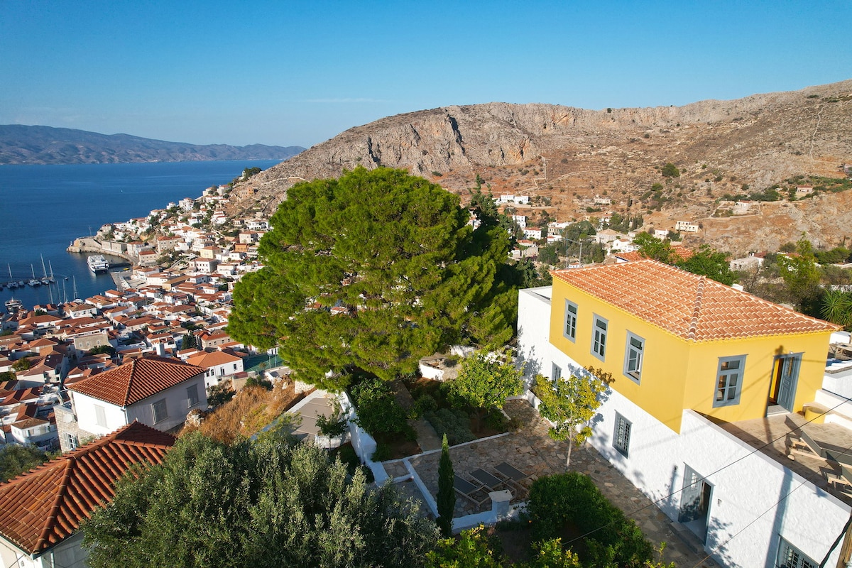 The image captures the villa situated on a hillside, featuring a large yellow building with a tiled roof. A lush green tree stands prominently in the foreground, providing a contrast to the rocky terrain and the scenic coastal view of Hydra and the Saronic Gulf in the background.