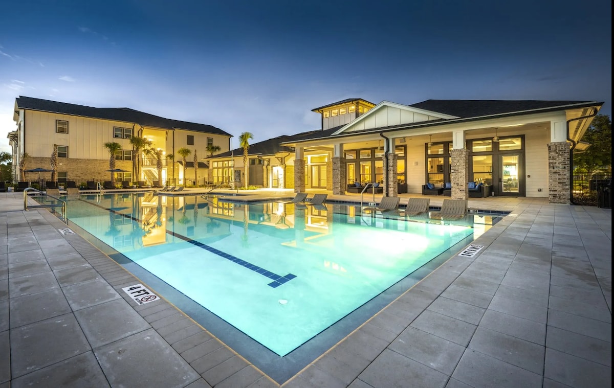 A resort-style pool area is featured, surrounded by modern lounge chairs. Large windows from nearby buildings reflect the evening light, while palm trees provide a tropical ambiance. The pool is well-lit, enhancing the inviting atmosphere of the outdoor space.