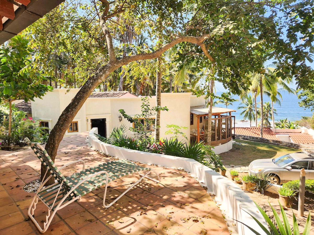 A spacious outdoor area features a relaxing lounge chair positioned beneath a leafy tree. The view reveals a tranquil beachfront setting, complemented by palm trees and vibrant greenery. A separate structure is visible in the background, harmonizing with the natural surroundings.