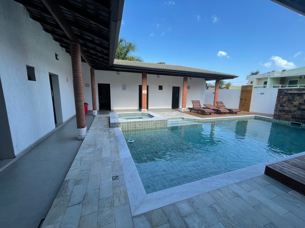 A sparkling swimming pool is surrounded by a sleek stone deck featuring two lounge chairs for relaxation. A small adjacent hot tub is visible, and changing rooms are located nearby, all under a light blue sky.