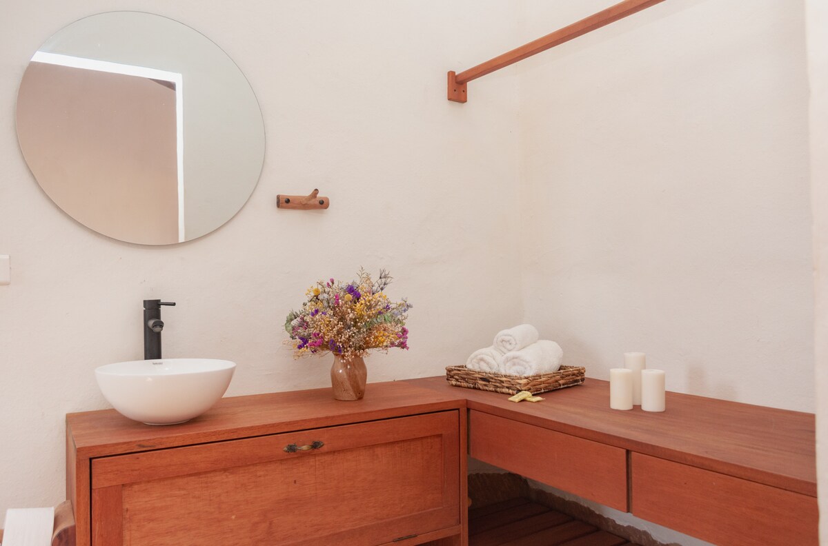 A modern bathroom features a round mirror above a wooden vanity. A white vessel sink is positioned beside a bouquet of dried flowers in a vase. Fresh towels are neatly arranged on a wooden shelf, complemented by soft candles on the countertop.