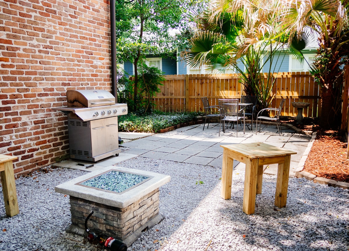 An outdoor patio area features a gas BBQ grill, stone fire pit, and wooden tables surrounded by greenery. Smooth stone slabs form the flooring, and a wooden fence provides privacy, enhancing the tranquil setting for gatherings.