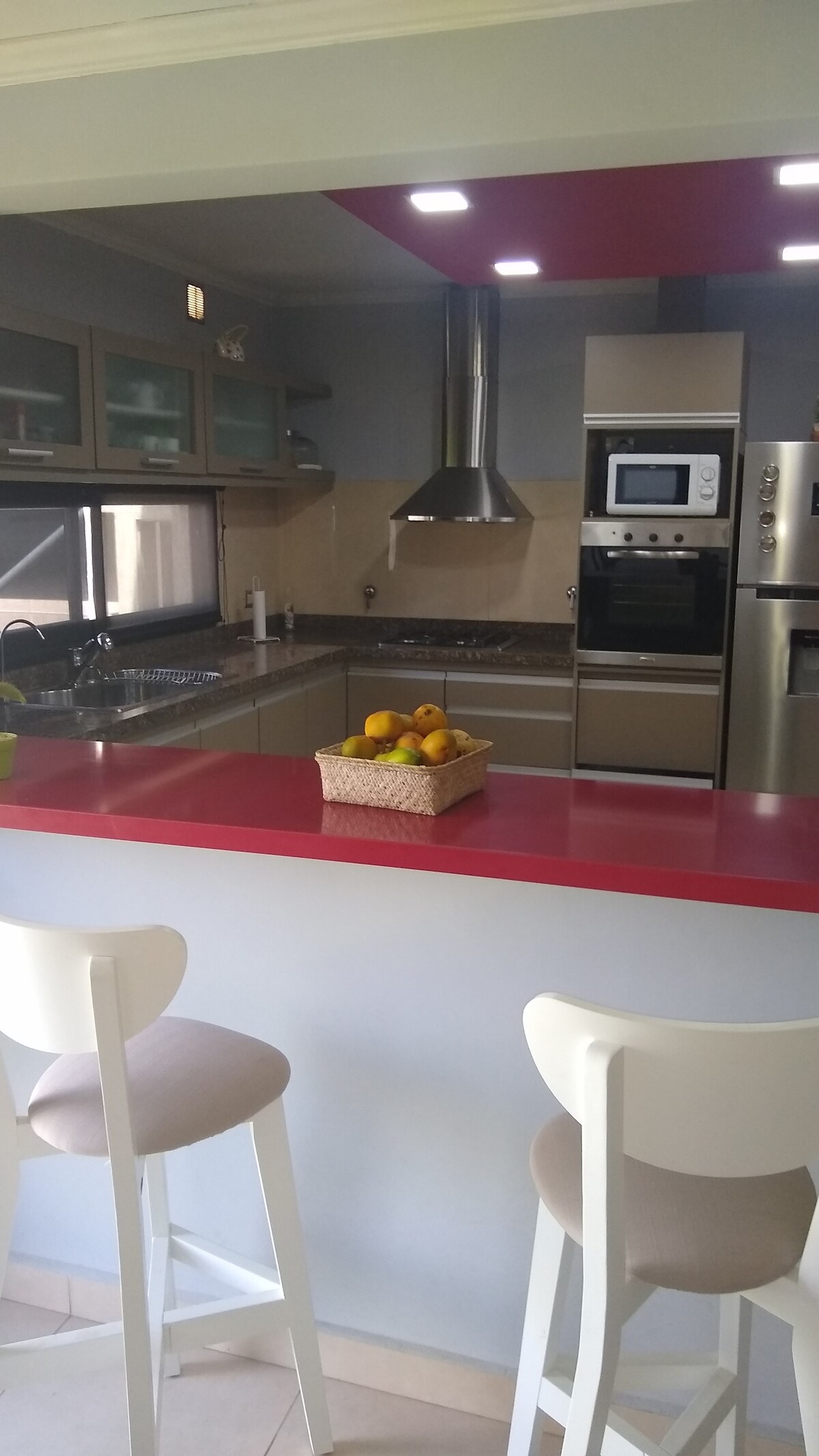 A well-appointed kitchen is showcased, featuring sleek cabinetry and a modern color palette. A red bar counter is accented by a basket of fresh oranges, while light fixtures illuminate the space. Two high-backed stools provide seating at the counter, enhancing the kitchen's inviting layout.