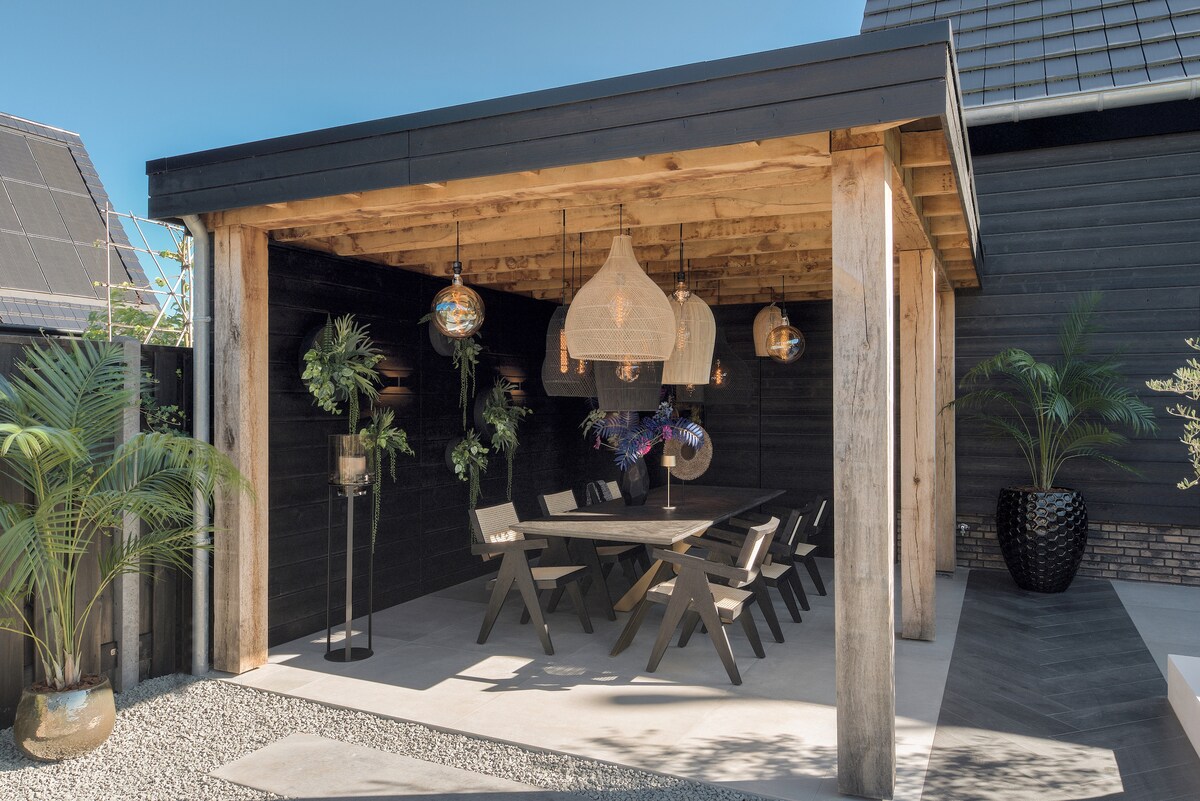 An outdoor dining area features a substantial black wooden table surrounded by matching benches. The space is illuminated by decorative hanging lamps and is adorned with potted plants, creating a cozy setting under a stylish wooden structure.