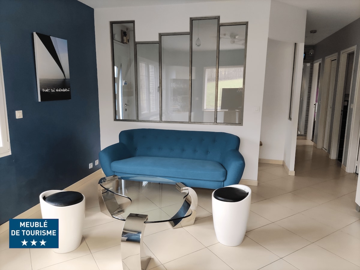 A modern living area features a sleek blue sofa positioned in front of a glass coffee table. Geometric partitions create visual interest while natural light floods in through large windows, offering a glimpse of the outdoor greenery.