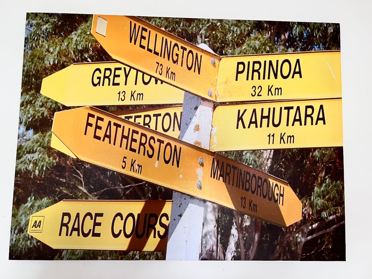 A directional signpost is presented with multiple yellow arrows pointing toward various destinations. Each arrow displays the name of a location, accompanied by the distance in kilometers. The background features foliage, enhancing visibility of the sign.