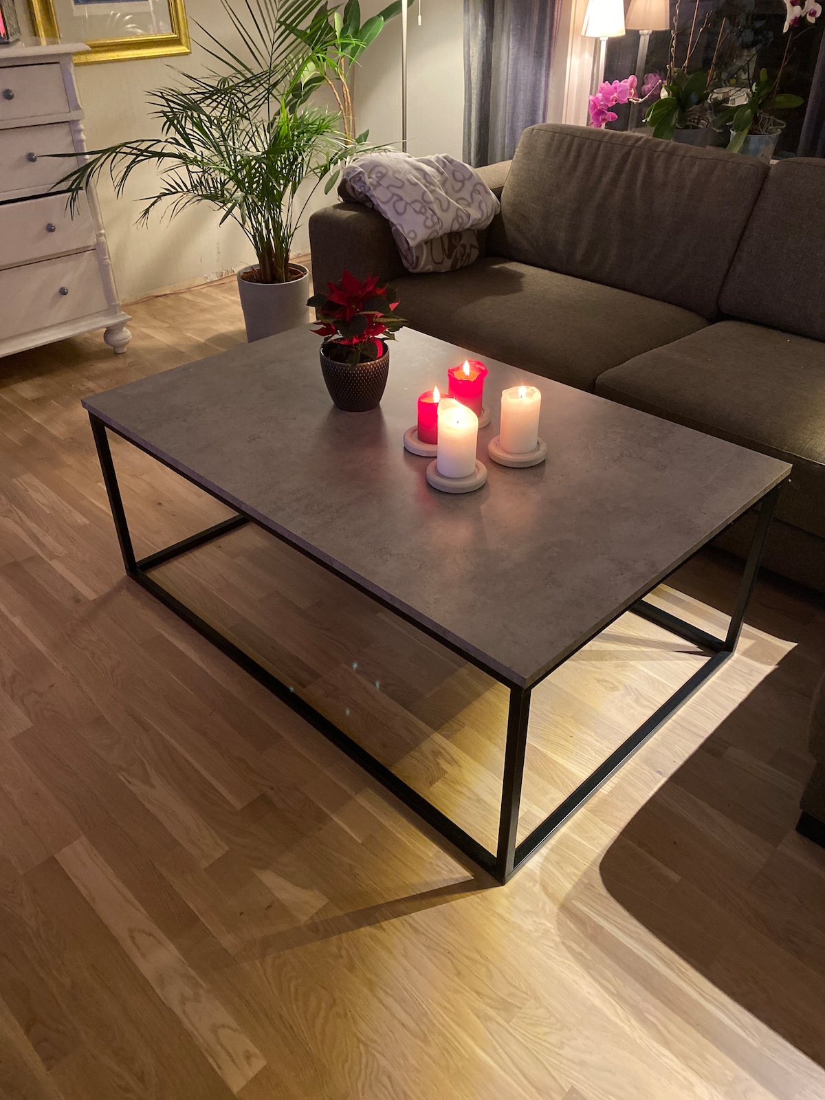 A modern coffee table with a concrete surface is positioned in front of a comfortable sofa. Soft-lit candles in varying heights create a warm ambiance, resting on a white plate. A decorative plant adds a touch of greenery to the space, enhancing the welcoming environment.