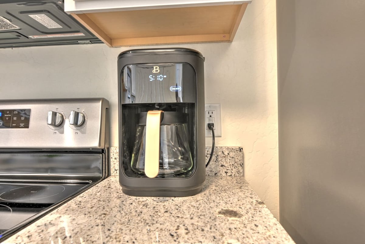 A modern coffee maker is positioned on a granite countertop, featuring a sleek design and digital display. Nearby, a stainless-steel stove adds a contemporary touch to the kitchen space, which is well-lit and designed for convenience.