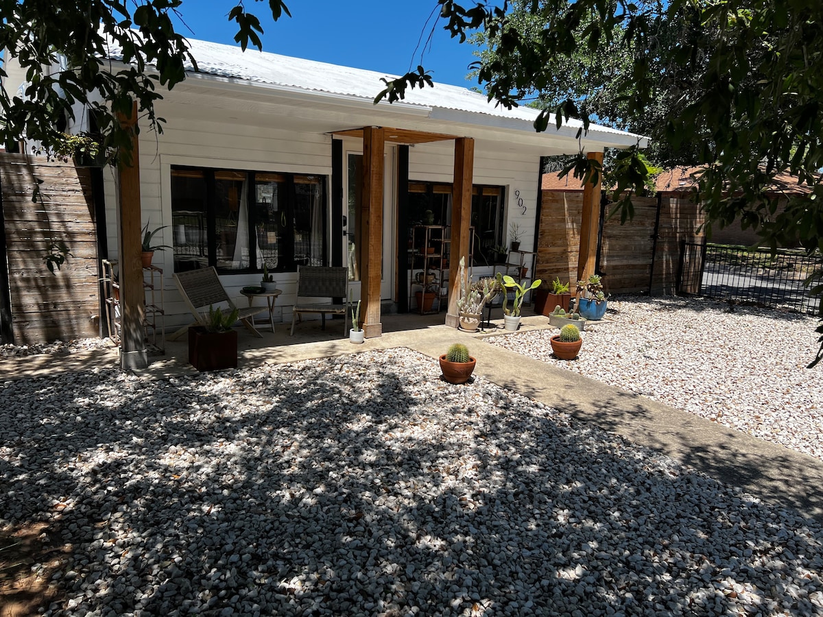 The exterior features a welcoming front porch with two chairs and small tables, surrounded by various potted plants. Sunlight filters through the trees, casting dappled shadows on the white gravel pathway that leads to the entrance.