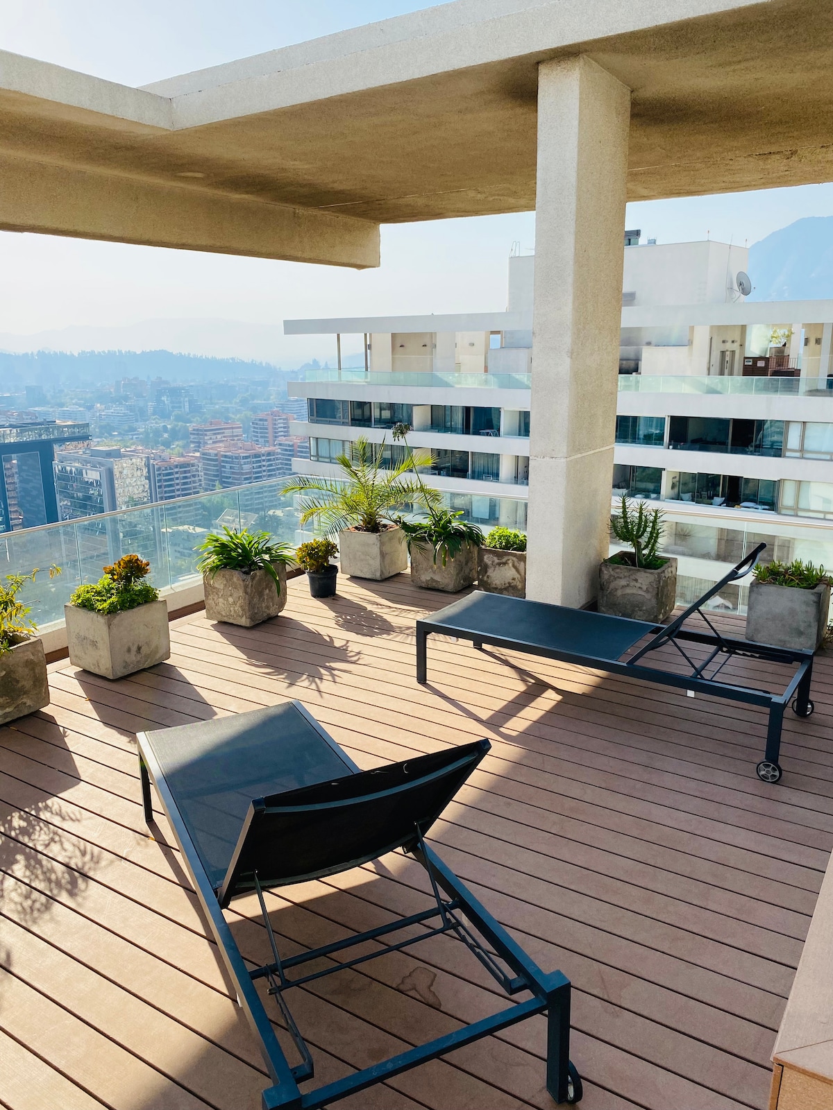 A furnished terrace is presented, featuring two lounge chairs arranged on a wooden deck. Potted plants are placed along the perimeter, and expansive views of the cityscape and distant mountains are visible beyond the railing.