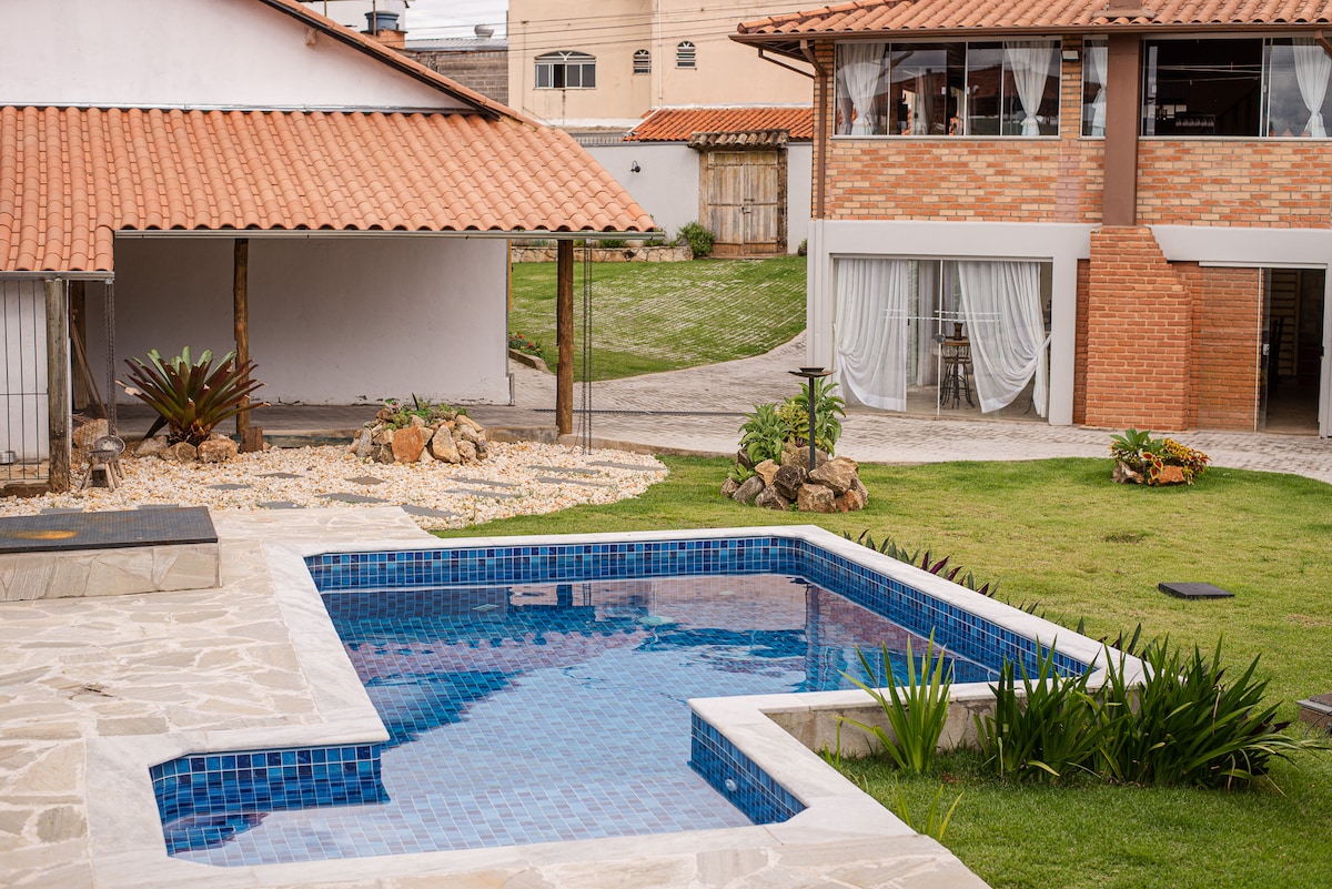 A rectangular pool is set in a tiled area, surrounded by manicured green grass and decorative stones. A nearby garden features various plants, adding a natural touch. In the background, a rustic building with a terracotta roof and large windows is visible, creating a serene outdoor environment.