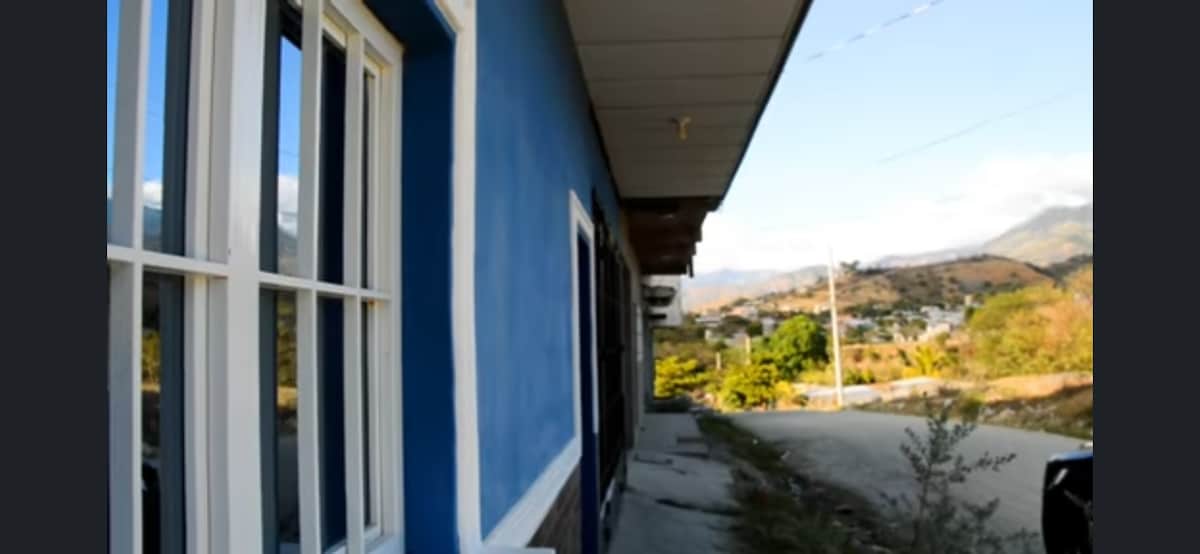 A side view of the property reveals a vibrant blue exterior complemented by white trim. Large windows allow natural light to brighten the space, while a pathway leads towards greenery and distant mountains, showcasing the peaceful surroundings.