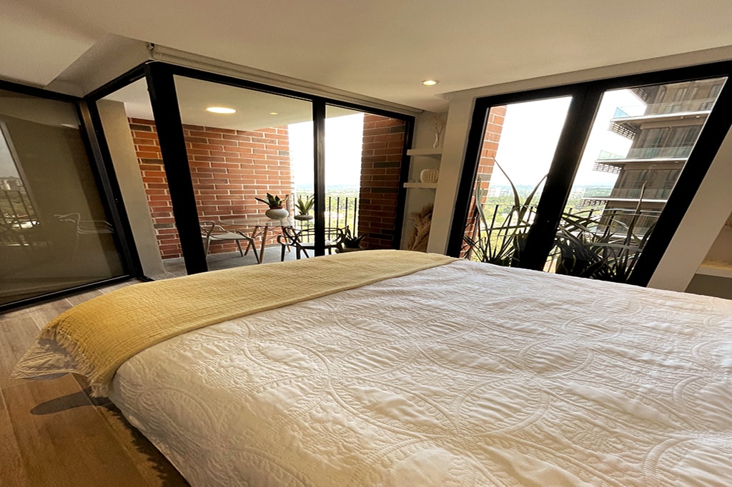 A spacious bedroom features a light-colored quilted bedspread draped over a large bed. Floor-to-ceiling windows offer views of a balcony with garden furniture and surrounding greenery, allowing natural light to fill the room.