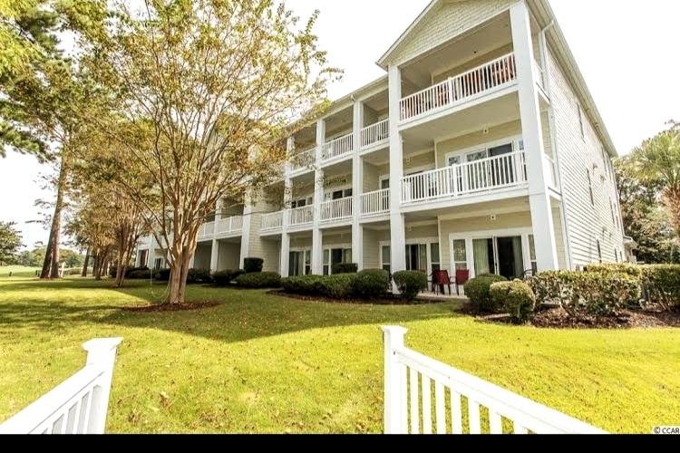 The exterior of a three-story condo building is presented, featuring multiple balconies with white railings. Lush green grass and neatly trimmed bushes surround the property, providing a well-maintained landscape. Tall trees contribute to the natural setting, enhancing the overall appeal of the building.