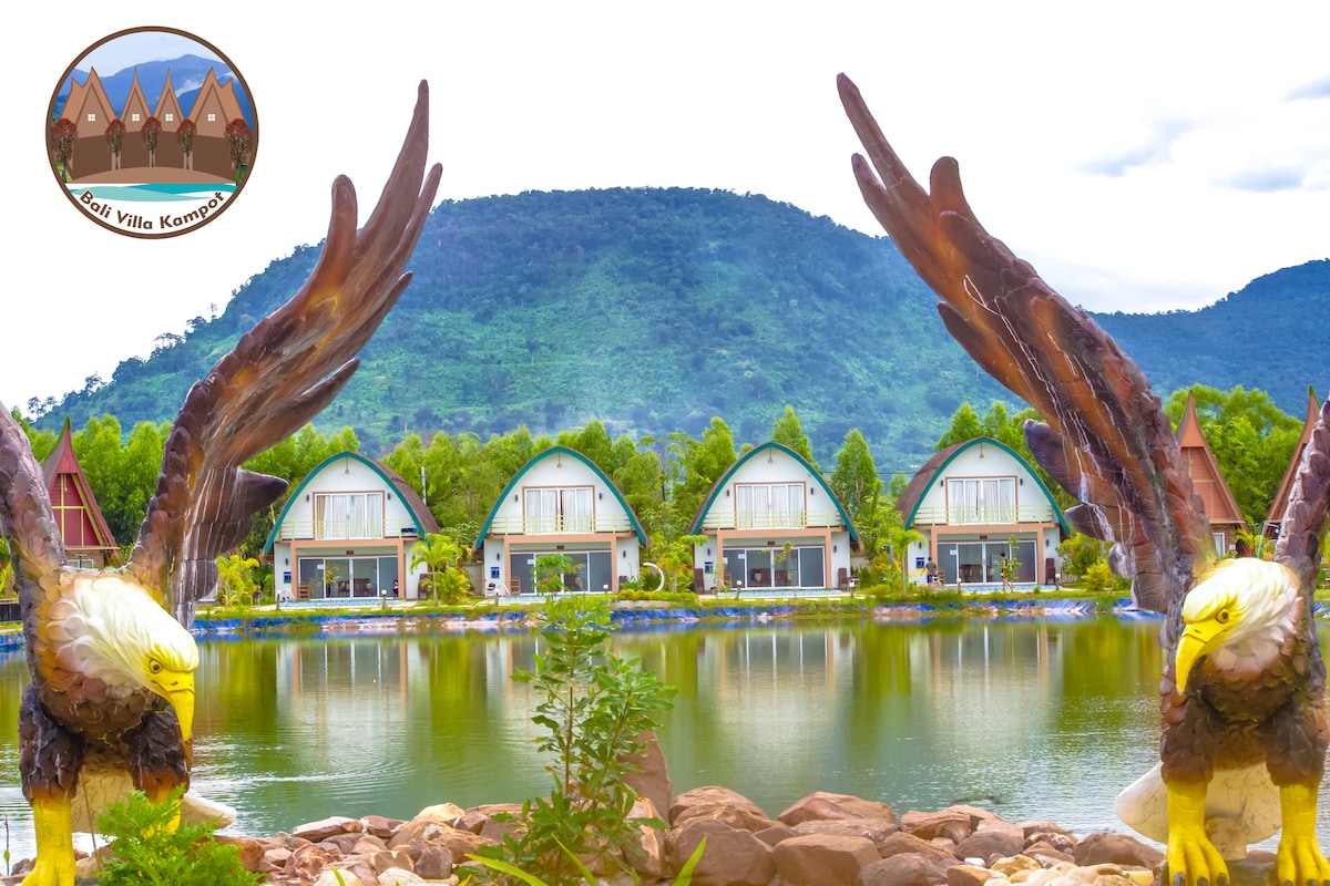 A serene view showcases the villa complex reflected in the calm waters of a man-made pond. Two large sculptures of eagles frame the entrance, while lush greenery surrounds the buildings, which feature slopes roofs and large windows that provide a modern aesthetic.