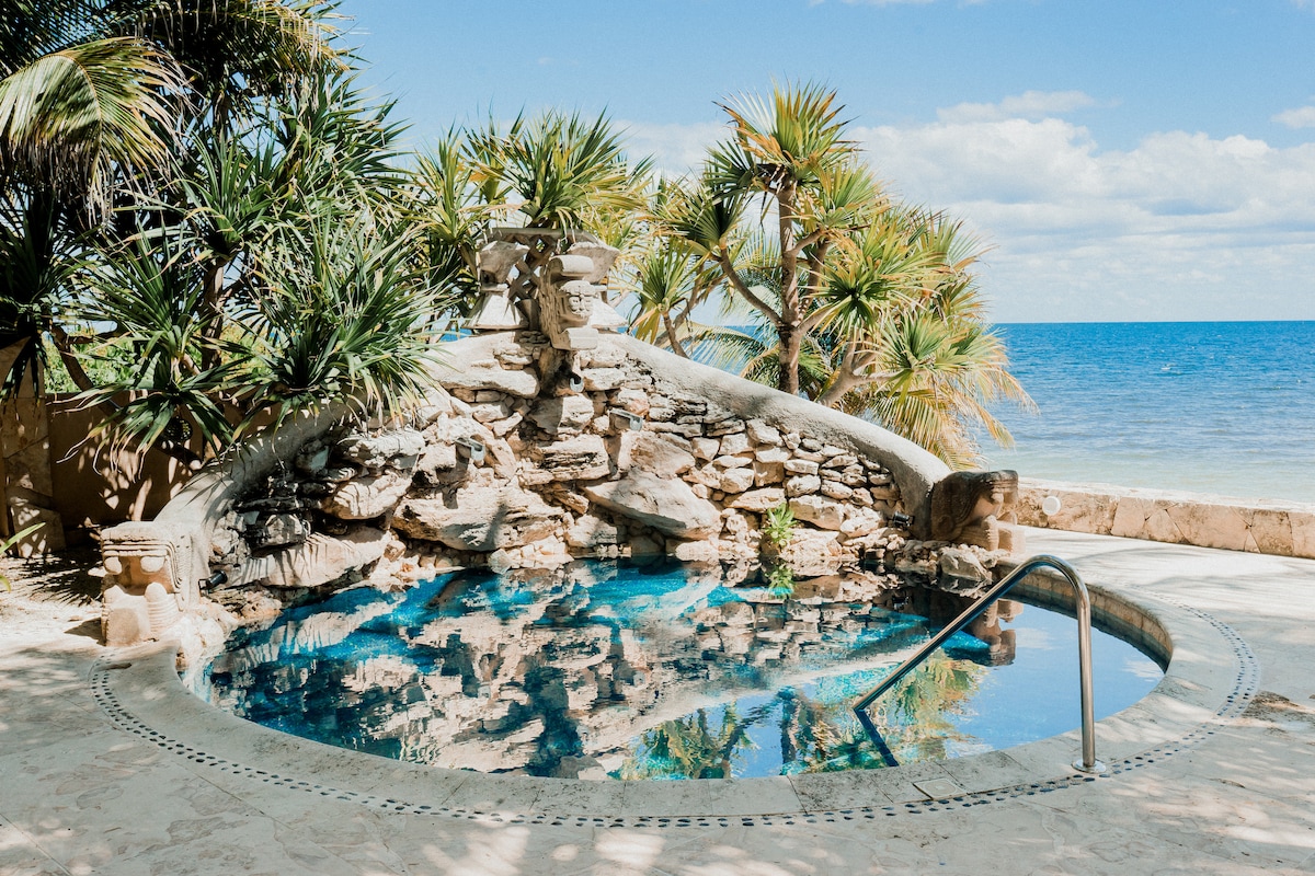A private hot tub is set against a backdrop of lush tropical plants and the ocean's edge. The clear water reflects the sky, creating a serene atmosphere. Natural rock features surround the tub, offering a seamless blend with the surrounding environment.